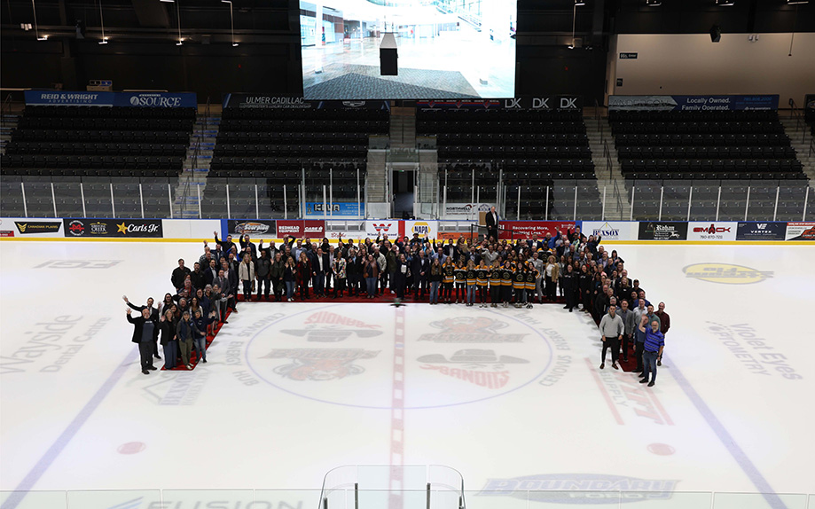 A large crowd stands in a U-shaped formation on the centre ice of a NHL-sized hockey arena. The photo is taking from above, presumably at the top of the spectator area. 