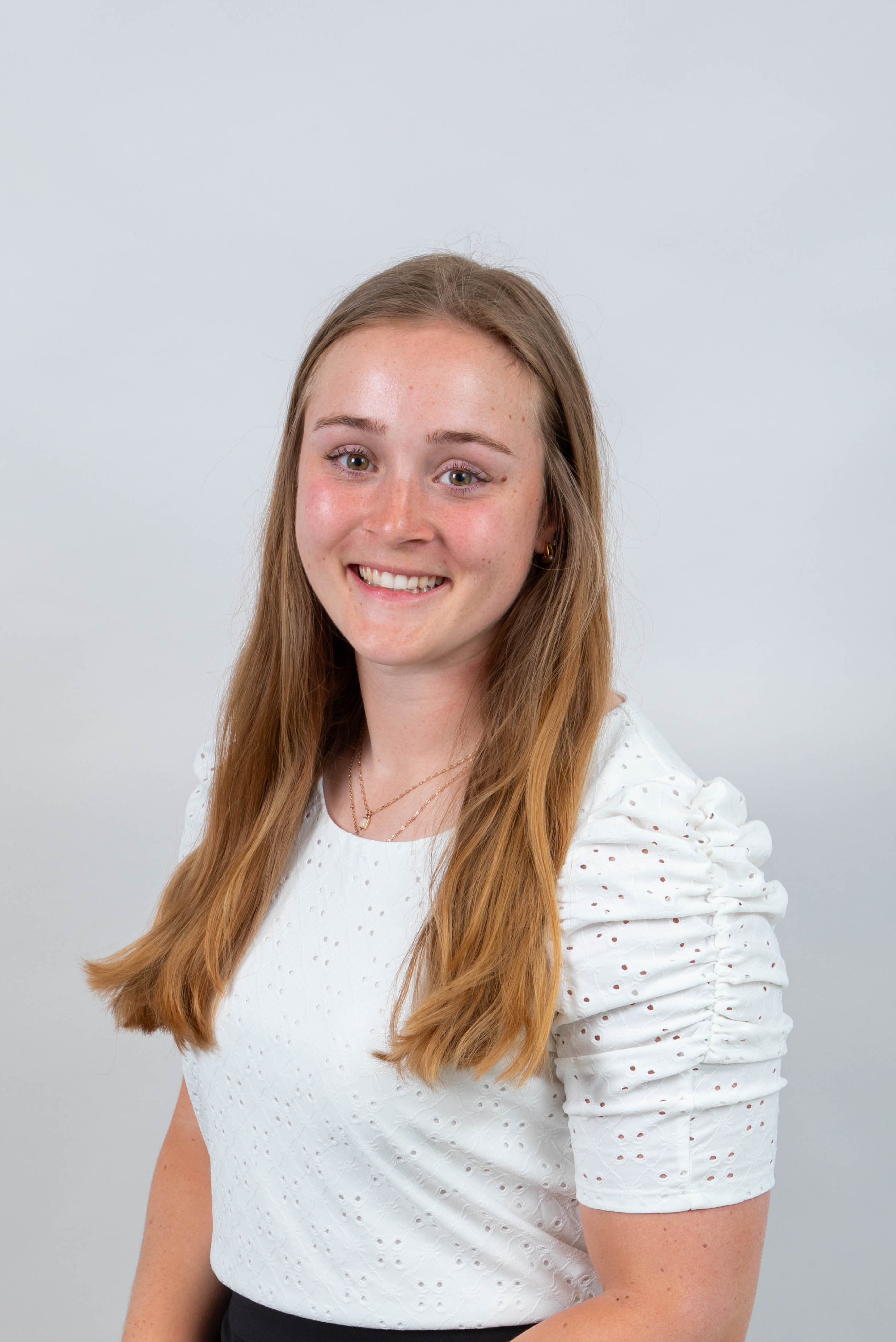 A professional headshot of a young woman. She has long, light-brown hair and is wearing a white, short-sleeve blouse. The background is light grey. 