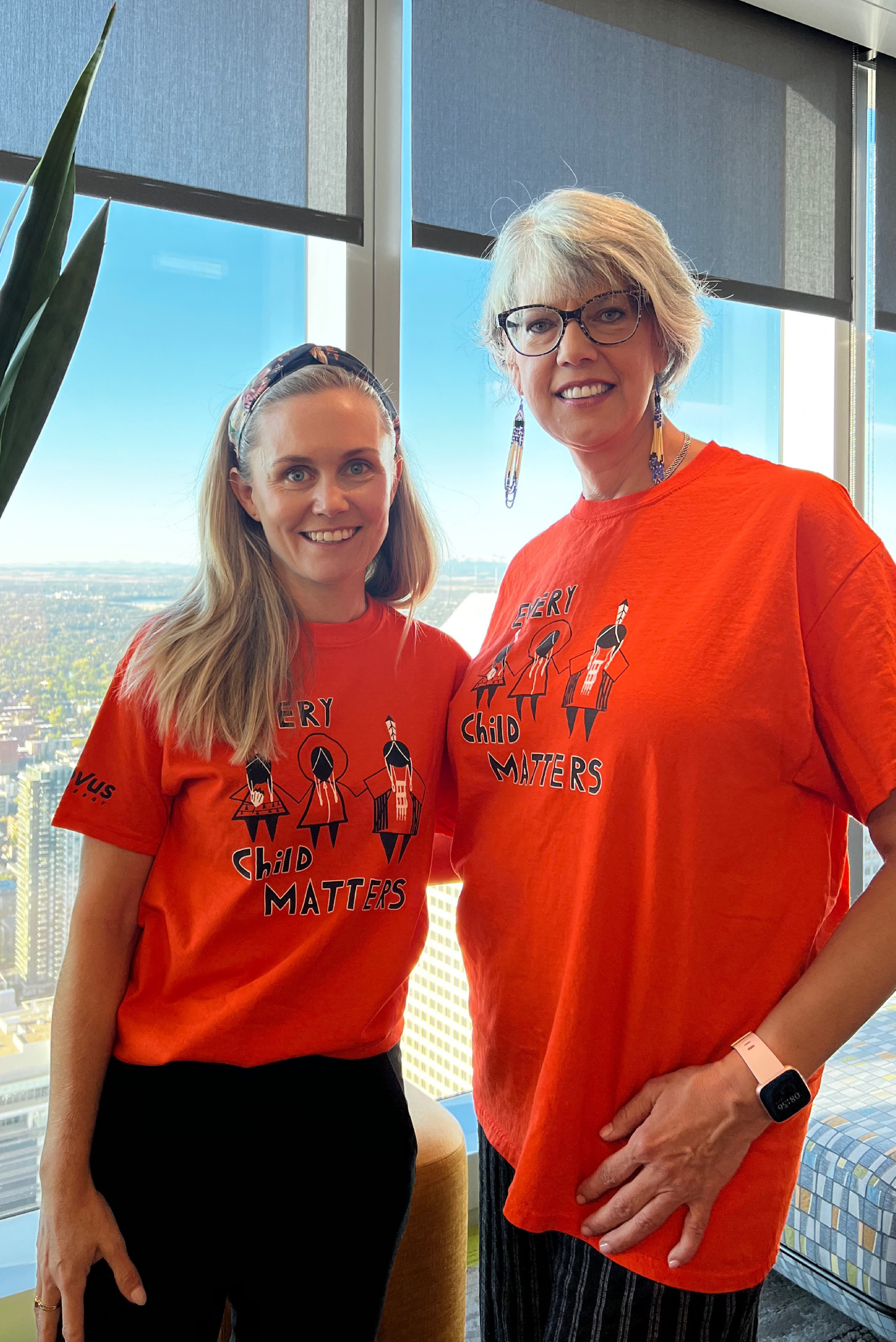 Two women are standing side-by-side in front of a window. They are wearing orange t-shirts that say "Every Child Matters." They are smiling at the camera. 