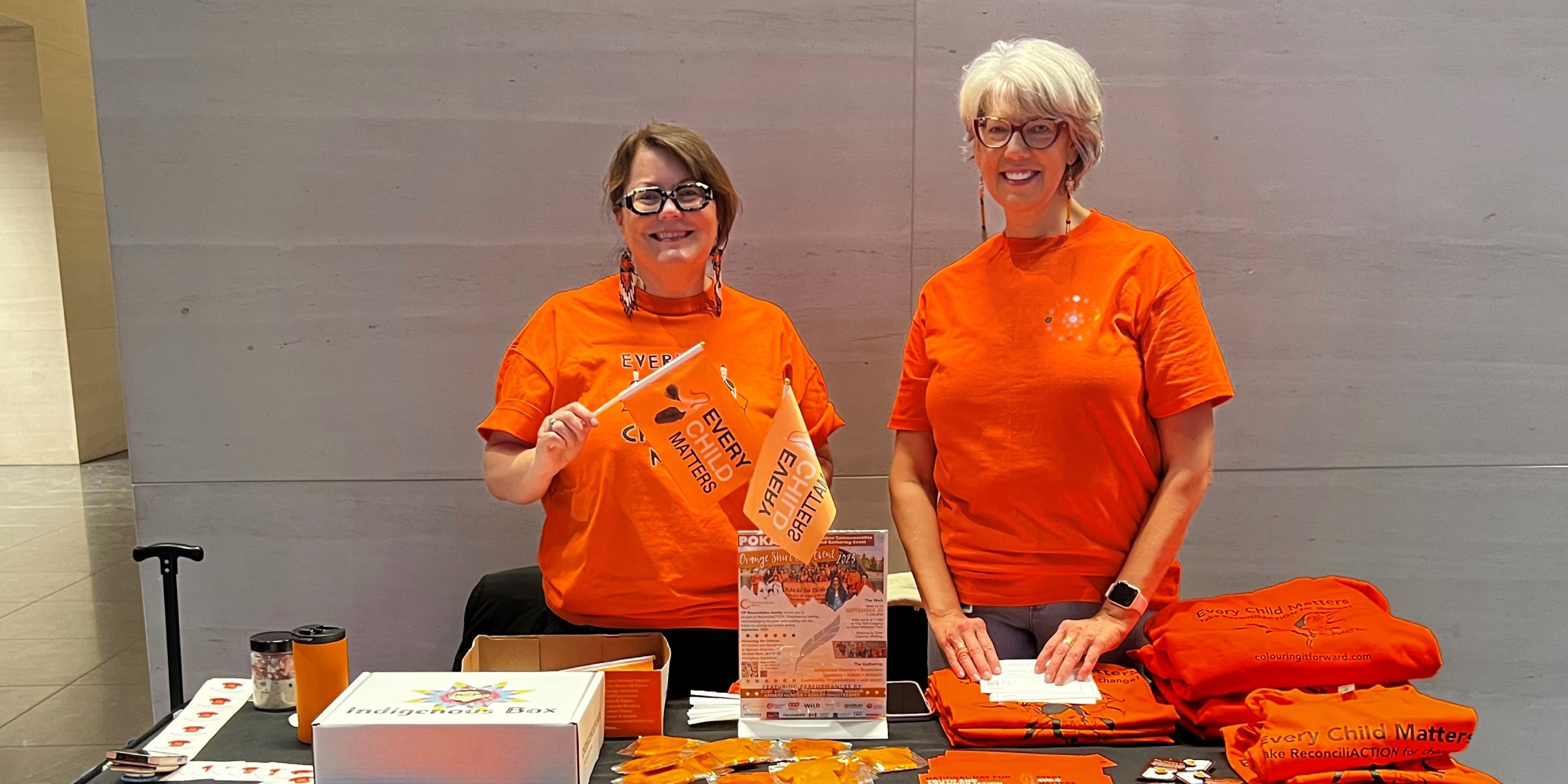 Two women are standing behind a table in a building lobby. The table has educational and orange materials in honour of Orange Shirt Day. They are both wearing orange shirts. 