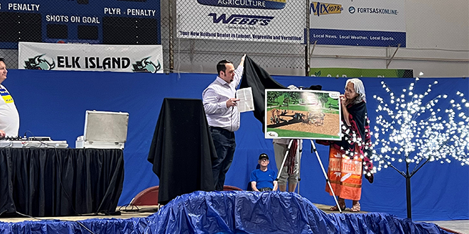 Two people are standing on a stage. They are removing a cover from a poster. The poster shows a digital rendering of an outdoor playground. 