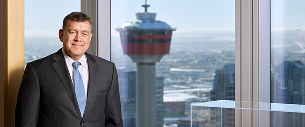 Cenovus President & Chief Executive Officer Jon McKenzie stands in an office while smiling. The Calgary Tower can be seen through the window behind Jon.